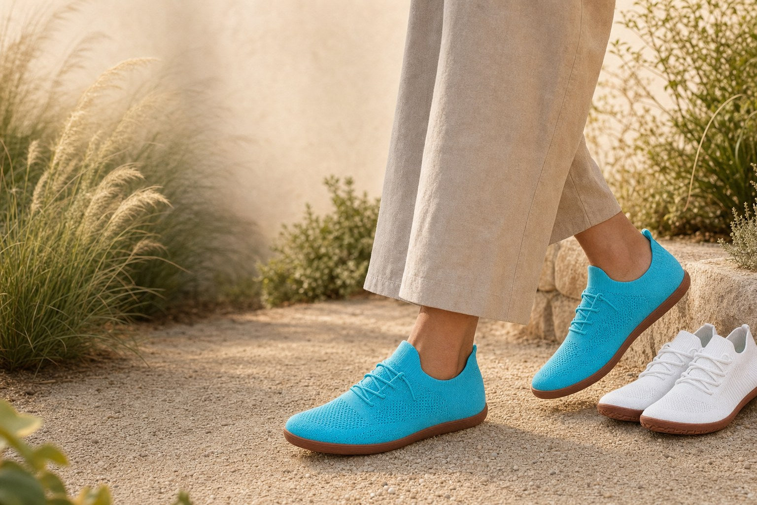 Woman in her fifties walking a sunlit garden path wearing Aqua Blue OnBarefoot barefoot sneakers, a white pair beside her